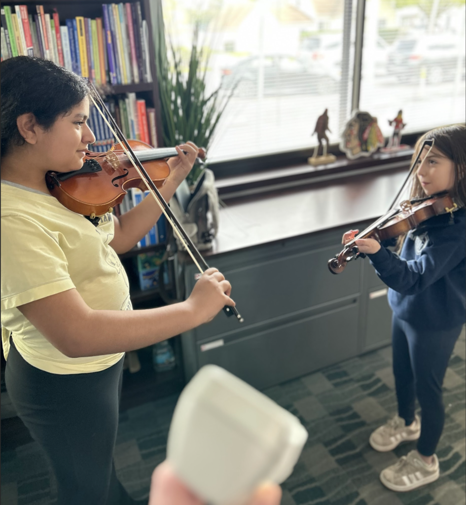 Two girls stand in a bright room, facing each other while playing violins. The girl on the left wears a yellow t-shirt and black pants, holding her violin up to her chin. The smaller girl on the right wears a navy sweatshirt and leggings, also holding her violin in a playing position. In the background, there is a bookshelf filled with books, a large window with blinds, and several small sports figurines on a ledge. The perspective is from slightly behind a white object in the foreground, creating a sense of being in the room during a lesson or practice session.