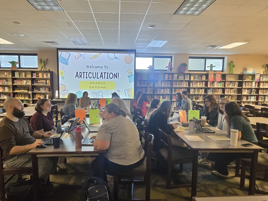 A group of teachers sit at tables in a school library collaborating in small groups, using laptops and papers. At the front of the room, a large screen displays “Welcome to Articulation!” with a schedule for ELA and Math sessions. Bookshelves line the walls, and the setting reflects a structured professional learning meeting.