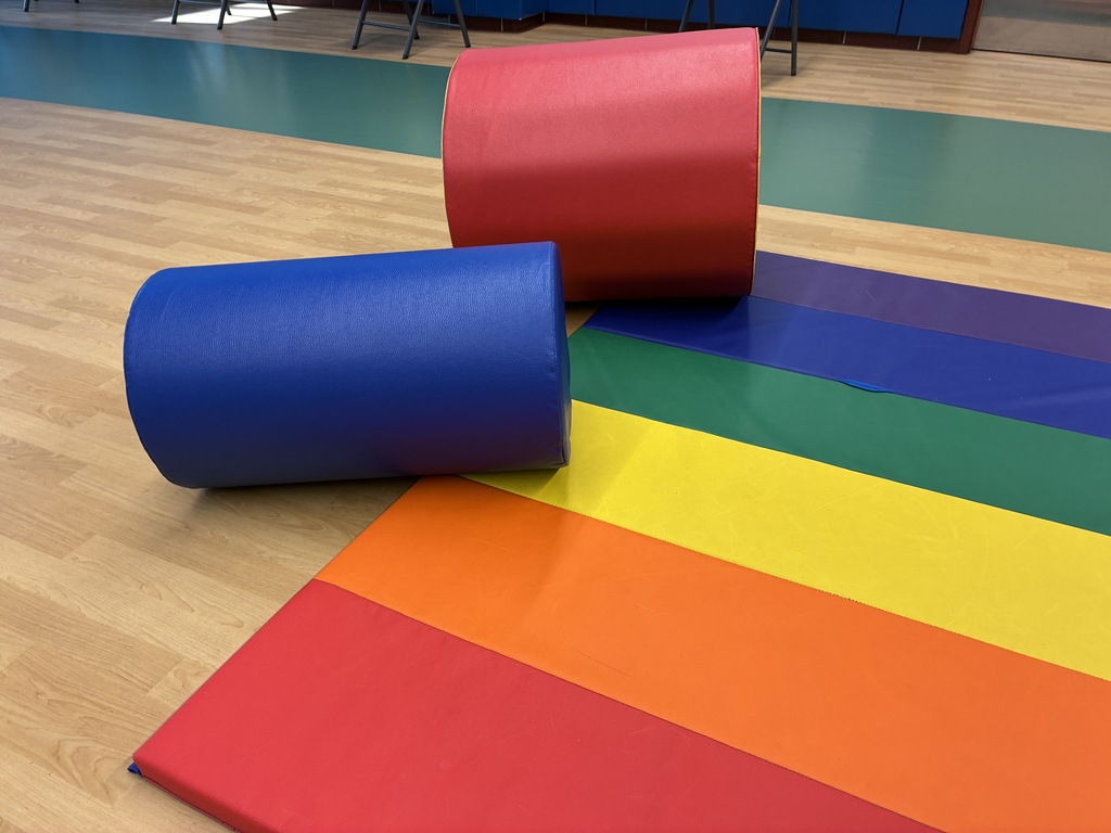 A red and a blue vinyl cylindrical bolster sit on a brightly colored rainbow floor mat in a school gym or therapy room with wood-laminate flooring.