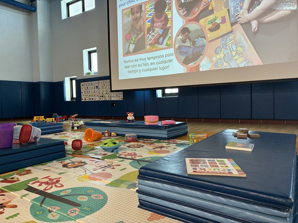 A brightly lit multipurpose room or gym set up for a parent-child workshop. Blue floor mats and colorful play mats are scattered with toys and children's books. In the background, a large projection screen displays a presentation in Spanish about the importance of reading to children at any age.