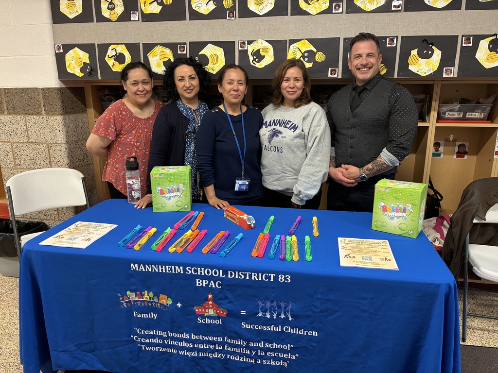 Five smiling adults stand behind a blue table for Mannheim School District 83 BPAC. The table features colorful bubble wands, flyers, and a tablecloth that reads "Family + School = Successful Children" in English, Spanish, and Polish. The background shows a classroom wall decorated with student artwork of bees.