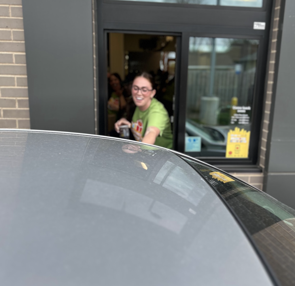 A woman with long dark hair and glasses, wearing a lime green t-shirt, leans out of a McDonald's drive-thru window with a bright smile. She is reaching toward a vehicle to hand over an item, while another person is visible smiling in the background inside the restaurant. The foreground shows the top of a dark gray car, reflecting the woman's face.