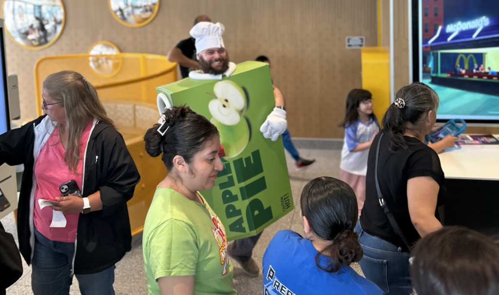 A medium shot inside a busy McDonald's restaurant shows a man in a chef's hat and a large, green Apple Pie mascot costume interacting with customers. The pie costume has a picture of an apple and the word "Pie." The chef has a broad smile. Other people are in the foreground, facing away from the camera, looking at the man. In the background, there's a McDonald's self-ordering kiosk and a video screen with the McDonald's logo. The area is filled with other people.