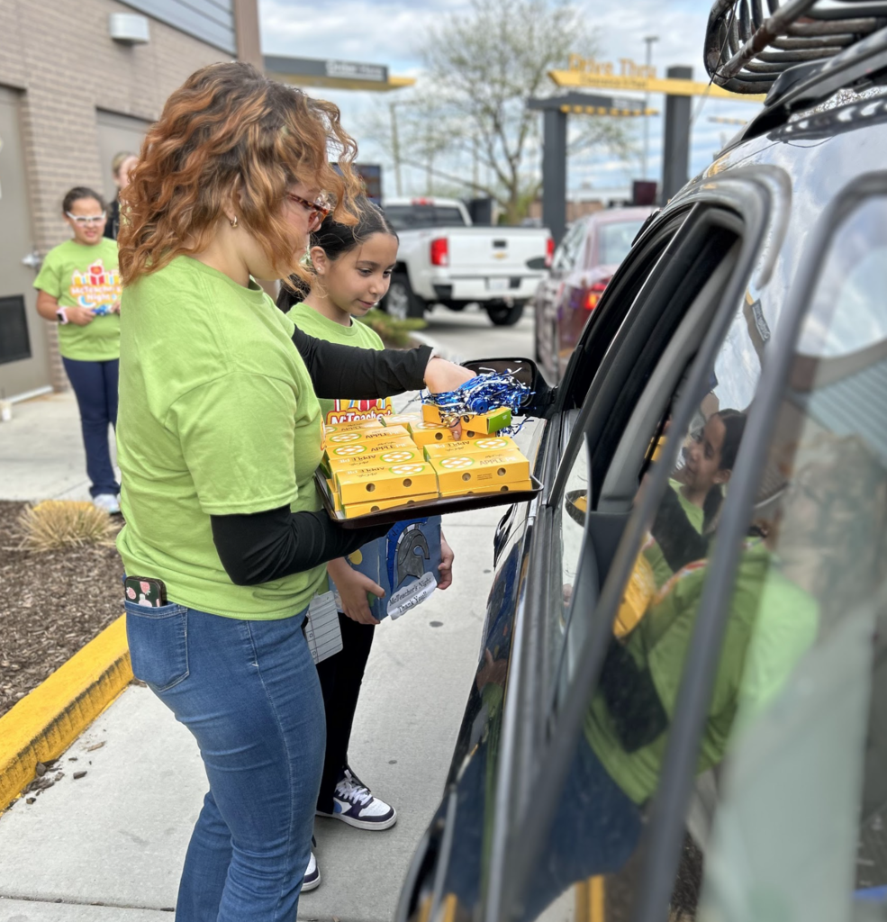 In a bustling drive-thru, a woman with vibrant, wavy red hair wears a light green "McTeacher's Night" t-shirt, jeans, and glasses, while carefully holding a tray filled with multiple yellow boxes. She leans towards a dark blue SUV, smiling at the car's window and handing one of the small, yellow boxes to a young girl inside.  Next to the woman, a young boy, also wearing a green "McTeacher's Night" t-shirt, stands attentively. Behind them, a young girl with a smile and glasses stands near a light grey building. The scene takes place during daytime at a drive-thru, featuring various vehicles and drive-thru signage in the background.