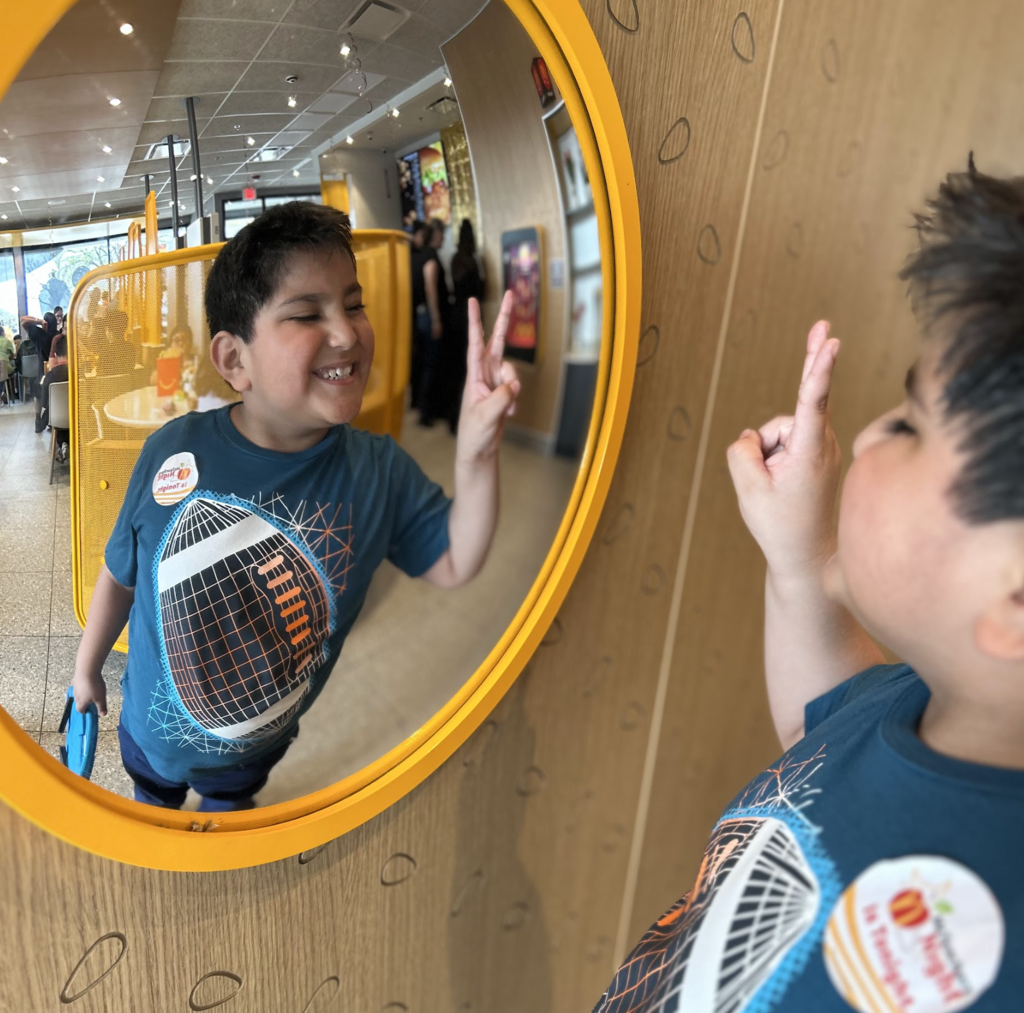 A close-up photograph of a young boy in a blue t-shirt with a graphic design and a round sticker. He is grinning and flashing a peace sign. He stands in front of a round, yellow-framed mirror, which reflects his smiling face and the peace sign back at him. The reflection also shows the inside of a brightly lit, modern fast-food restaurant.