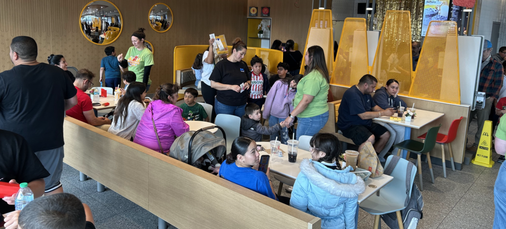 A crowded and lively McDonald's dining room is filled with families and children during a community event. Several women in lime green "McTeacher's Night" t-shirts are scattered throughout the room, interacting with guests and managing the busy atmosphere.  Families are seated at white tables and in booths with yellow mesh privacy screens, while others stand and talk in the aisles. In the background, circular mirrors on a tan patterned wall reflect the bustling scene. The restaurant is bright and modern, with a mix of casual seating and people of all ages enjoying the event.