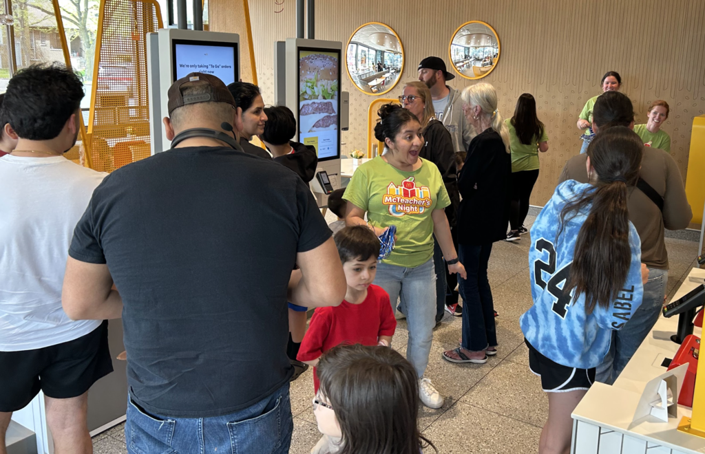 Inside a modern McDonald's restaurant, a diverse group of adults and children gather during a "McTeacher’s Night" event. In the center, a woman in a lime green event t-shirt and jeans looks toward a child with an excited, wide-mouthed expression.  Customers are seen from behind using digital ordering kiosks and standing in line, including a man in a black t-shirt and a girl in a blue tie-dye hoodie. In the background, other staff members in green shirts interact with guests near a brightly lit counter. The restaurant features clean, tan-colored walls with two circular mirrors reflecting the dining area.