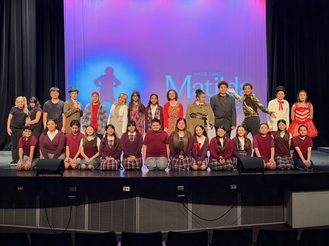 A large group of middle school students pose together on stage as the cast of *Matilda Jr.*, wearing colorful costumes representing various characters from the musical. Students are arranged in rows, smiling and celebrating their performance, with stage props and a theatrical backdrop visible behind them.