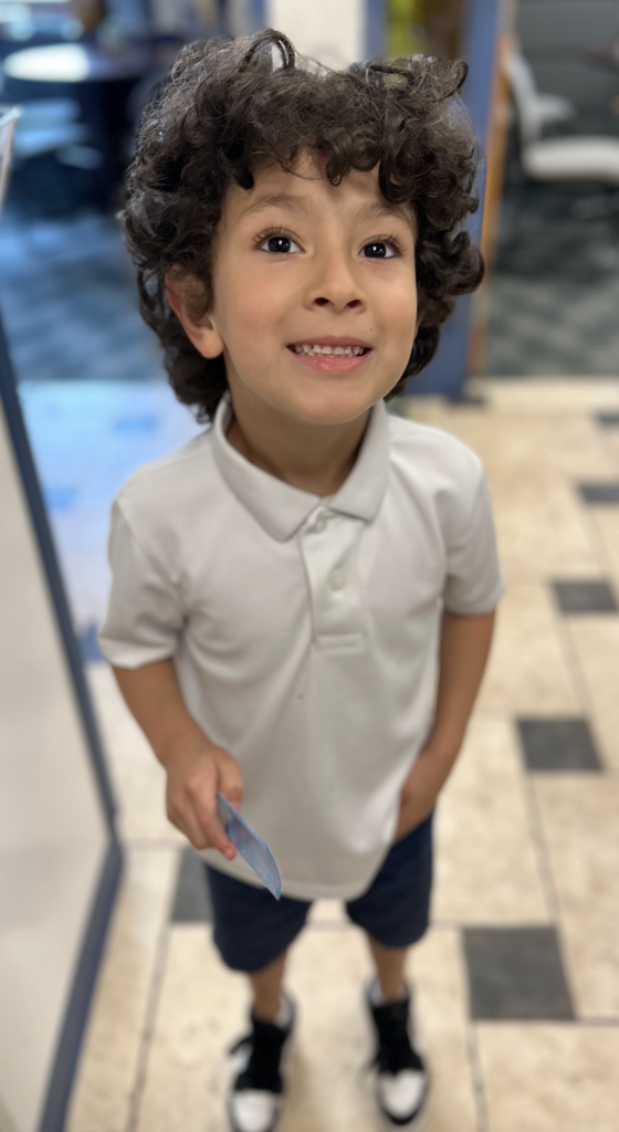 A high-angle, top-down photograph looking down at a young boy with dark, curly hair and brown eyes. He is wearing a white short-sleeved polo shirt and dark navy blue shorts, holding a small card in his left hand while looking up with a smiling expression. He is standing on a tiled floor with an irregular black and white pattern in a brightly lit indoor space.