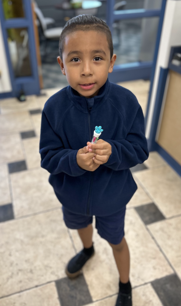 A young boy with short hair wearing a navy blue fleece jacket and matching shorts, standing on a black and white tiled floor. He is looking at the camera and holding a small, multi-colored pen with a blue cactus-shaped topper in both hands.