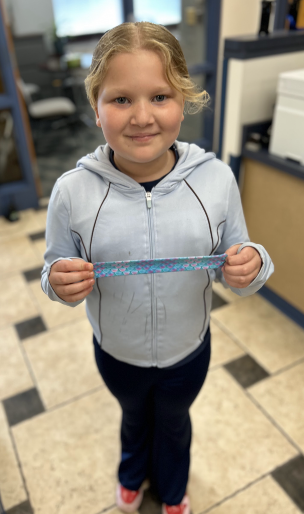 A high-angle, slightly blurred photograph of a young girl standing on a tiled floor. She is wearing a light blue zippered hoodie and dark blue flare pants, holding a mermaid-themed slap bracelet horizontally with both hands. She is smiling and looking directly at the camera. The background shows parts of a school or office setting.