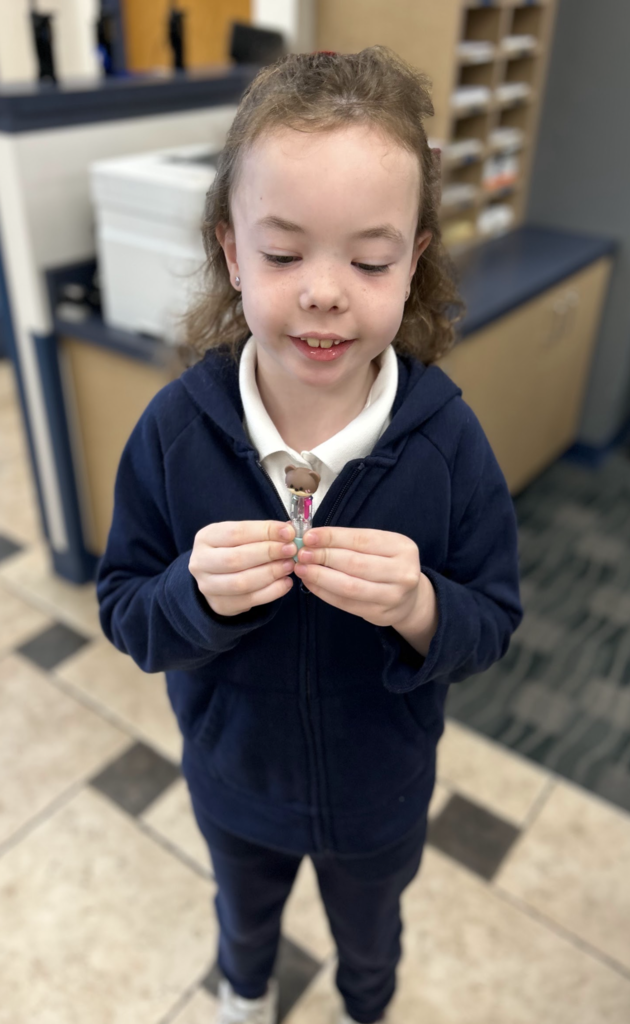 A young girl with light curly brown hair and a slight smile is holding and looking at a small, brown bear figurine with clear liquid inside it. She is wearing a navy blue zip-up hoodie over a white polo shirt, and navy pants. The background is a blurry office or school space with filing cubbies, a printer, and checkered floor tiles.