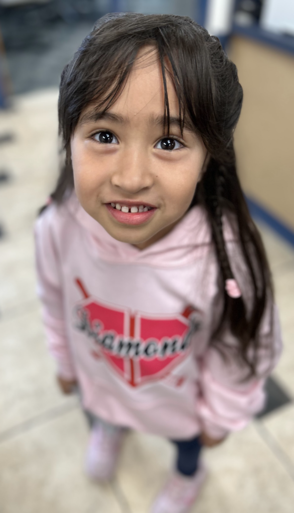 A high-angle, close-up portrait of a young girl with dark hair and bangs, smiling warmly at the camera. She is wearing a light pink hooded sweatshirt with a red diamond-shaped graphic on the front that includes the word "Diamond." Her hair is styled with small braids, and she is standing on a light-colored tiled floor.