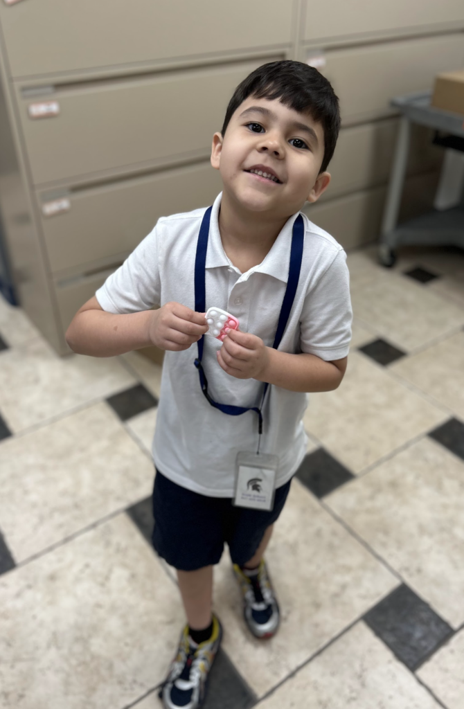 A young boy with dark hair and a smile stands in a room with tiled floors and filing cabinets. He is wearing a white polo shirt, dark shorts, a blue lanyard with an ID badge, and colorful sneakers. He holds a small, white and pink silicone "pop-it" fidget toy in his hands.