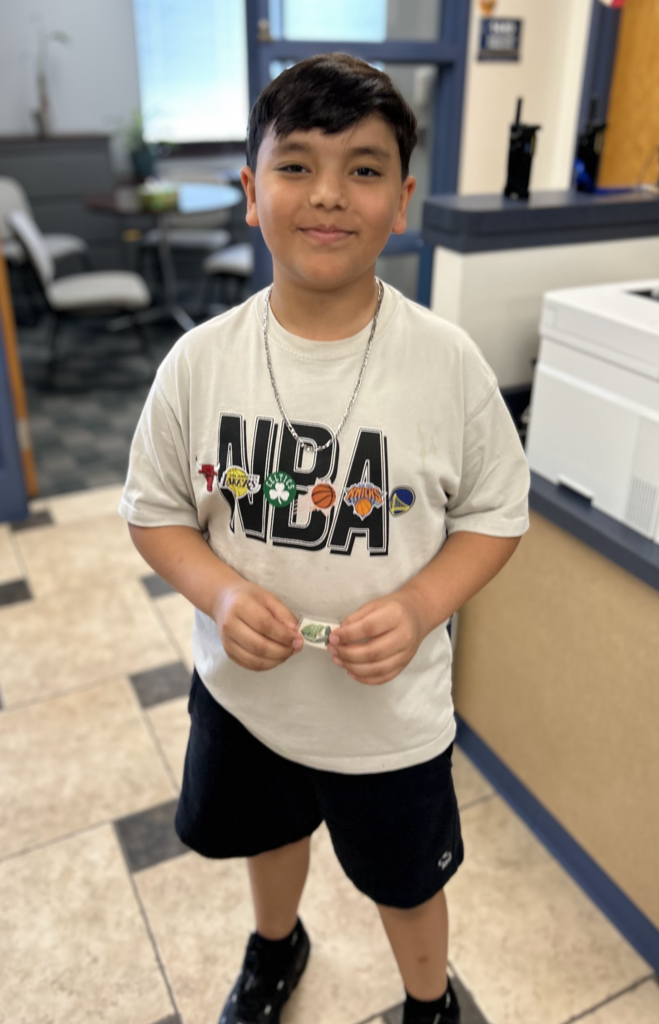 A medium shot of a young boy smiling indoors, wearing a white t-shirt with a large "NBA" logo featuring several professional basketball team emblems. He is wearing a silver chain, black shorts, and black sneakers, and is holding a small green sticker between his hands.