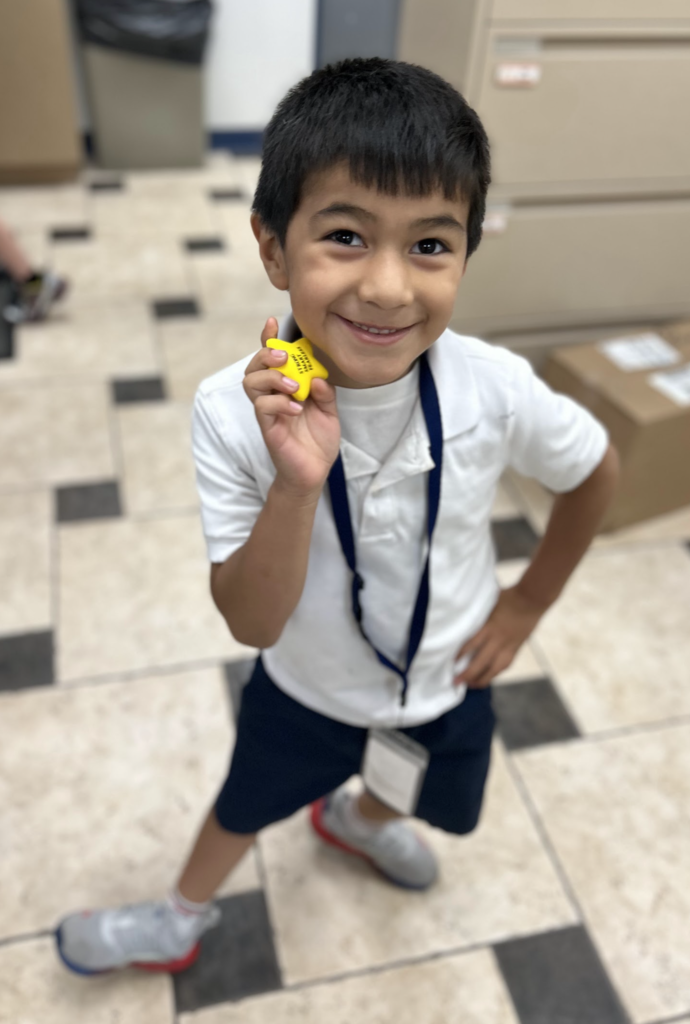 An image of a young boy with dark hair wearing a white polo shirt, blue shorts, a blue lanyard, and gray sneakers, standing in an office with a black and white tiled floor, smiling and holding a yellow star-shaped object next to his left ear.