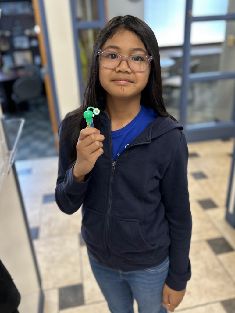 A medium shot of a young girl with long dark hair and glasses, wearing a navy blue zip-up hoodie over a blue shirt. She is standing in a room with tiled floors and holding up a small, green decorative pen or toy toward the camera.