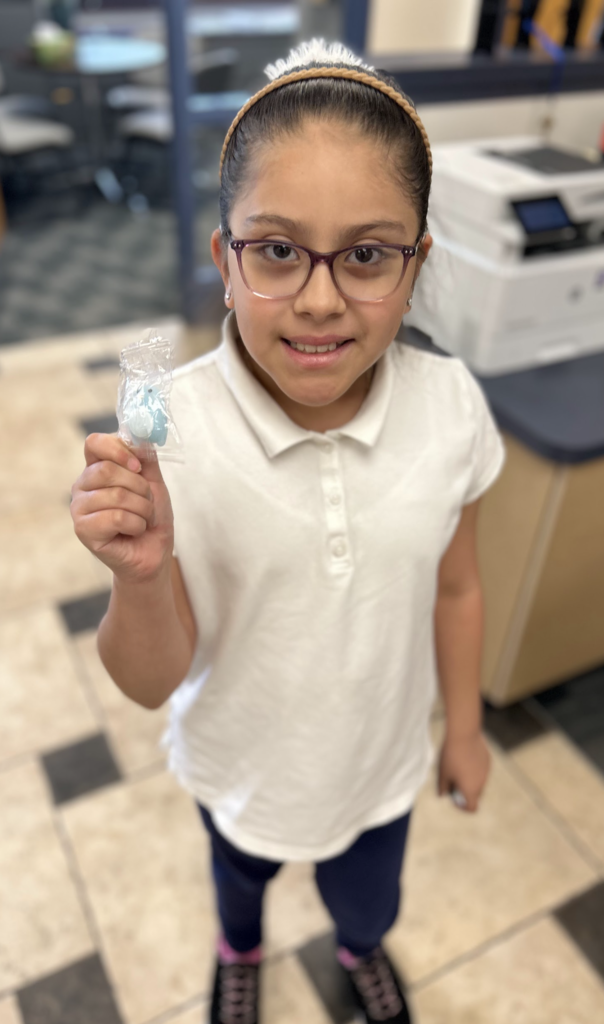A young girl with dark hair pulled back in a headband and wearing purple-rimmed glasses smiles at the camera. She is wearing a white short-sleeved polo shirt and dark pants. She holds up a small, light blue toy inside a clear plastic wrapper. The background features a tiled floor and a blurred office setting with a printer.