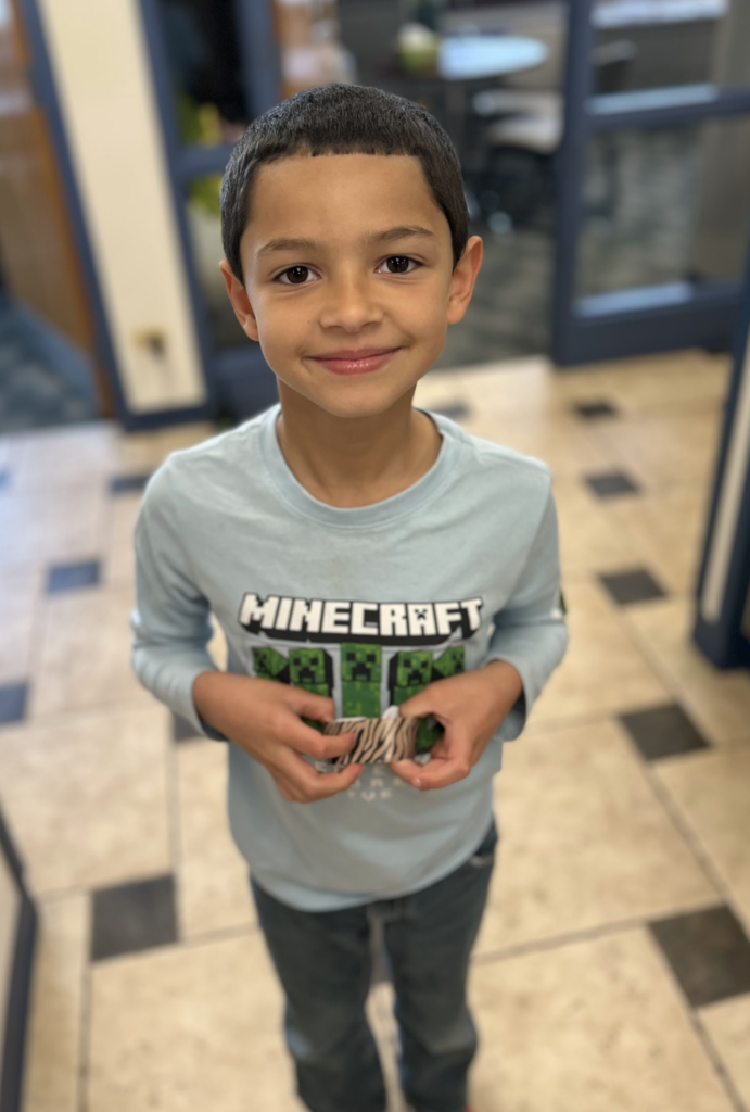 A young boy with a short haircut and a friendly smile stands in a school hallway. He is wearing a light blue long-sleeved Minecraft shirt and holding a small zebra-patterned item in both hands.