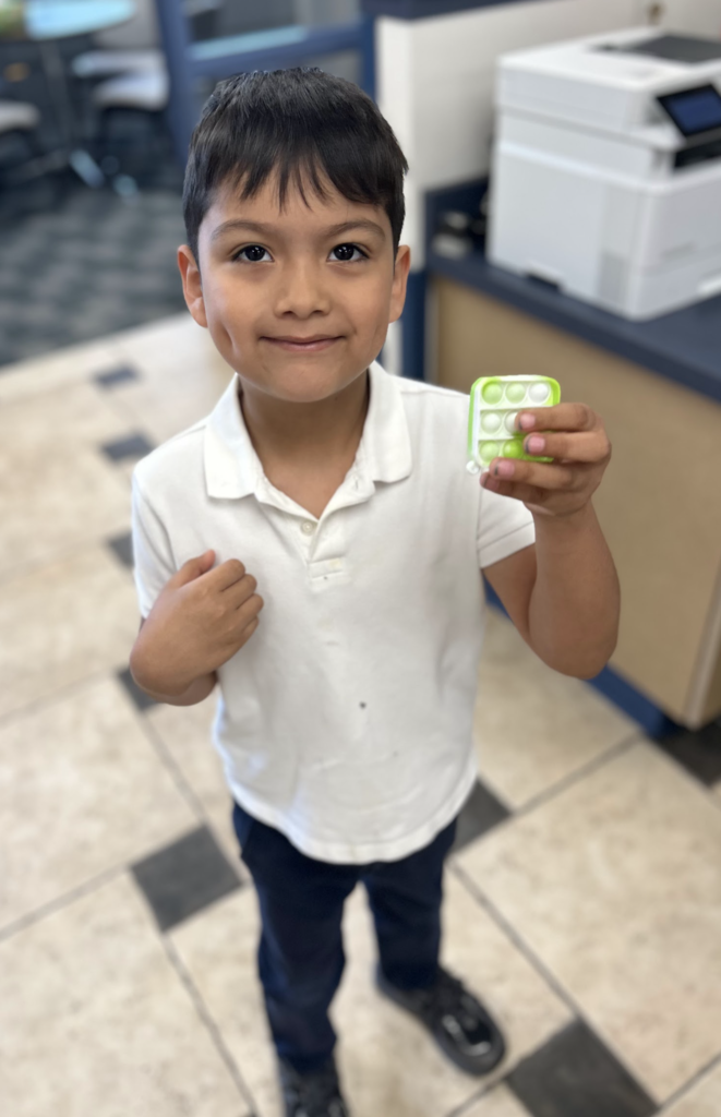 A young boy with dark hair and a slight smile stands in an office-like setting, wearing a white short-sleeved polo shirt and dark pants. He is holding up a small, square, lime-green "pop-it" fidget toy in his left hand. The background shows a light-colored tiled floor and a white office printer on a desk.