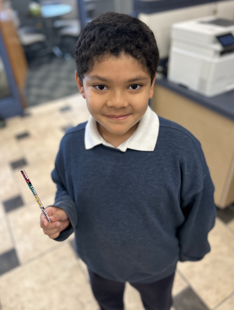 A smiling young boy with curly dark hair standing in a school office. He is wearing a navy blue sweater over a white collared shirt and is holding a colorful, patterned pencil.