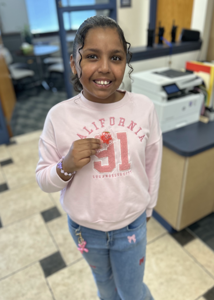 A smiling young girl in a light pink "California 91" sweatshirt and blue jeans stands in an office or classroom. She is holding up a small, red strawberry-shaped charm or candy in a plastic wrapper.