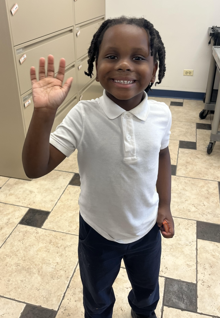 A young boy with dark hair in twists stands in a room, smiling broadly and waving with his right hand. He is wearing a white short-sleeved polo shirt and dark navy blue pants. He holds a small orange object in his left hand. The background includes tan filing cabinets and a light-colored tiled floor with dark accent squares.