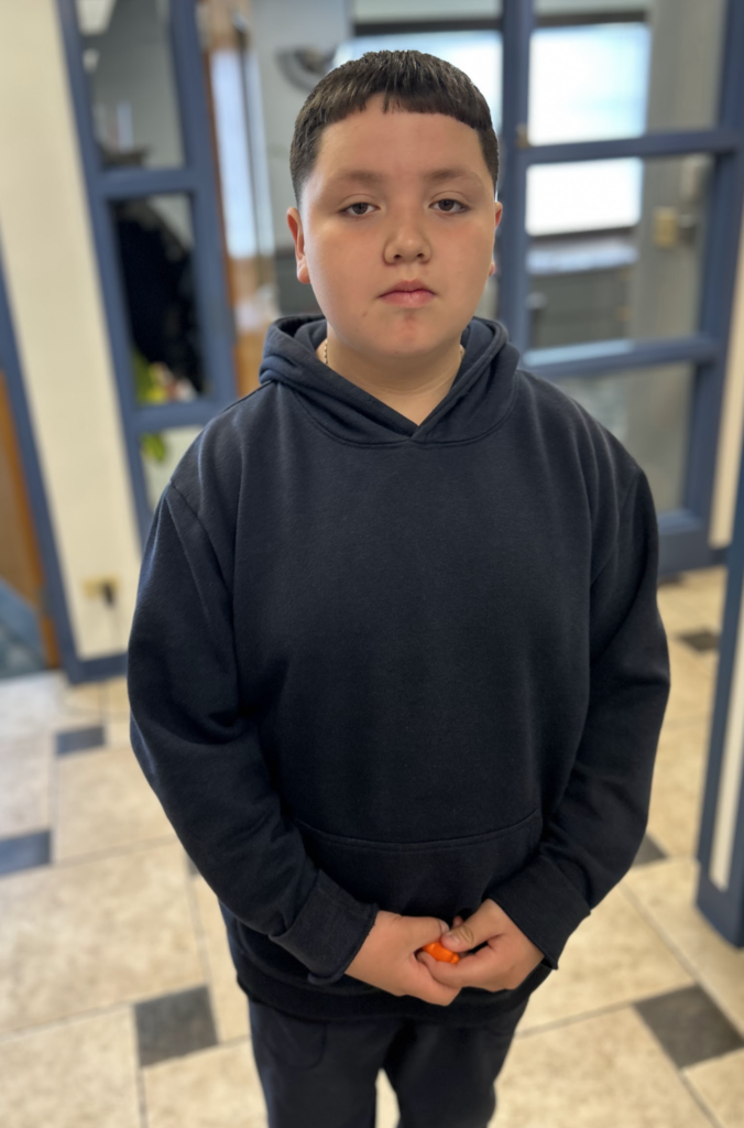 A medium shot of a young boy with dark hair and a short fringe standing in a room with blue-framed doors and a tiled floor. He is wearing a dark navy blue hoodie and dark pants, and he is holding a small orange object in his hands.