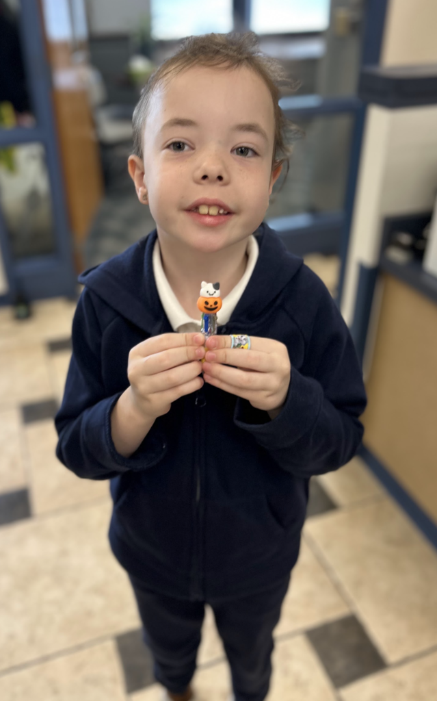 A smiling young boy in a navy blue zip-up hoodie stands in a school office. He is holding a small toy or pencil topper featuring a jack-o'-lantern and a white cat with both hands.