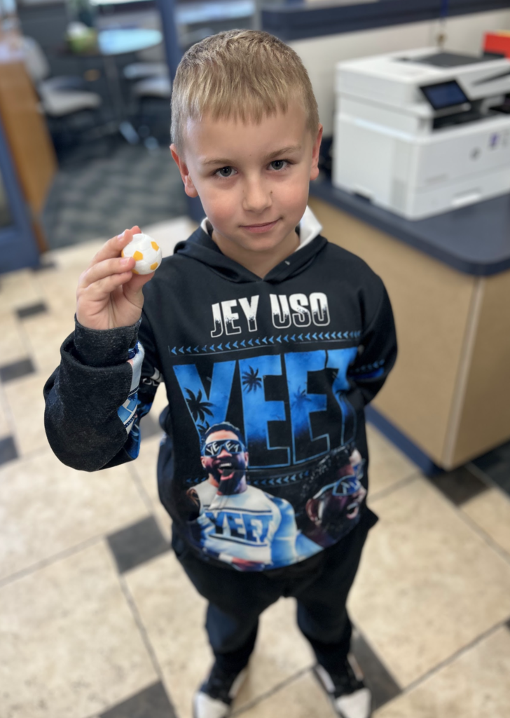 A young boy with short blonde hair stands in a school office, holding up a small white and orange soccer ball-shaped toy. He is wearing a black hoodie with "Jey Uso" and "Yeet" graphics on the front.