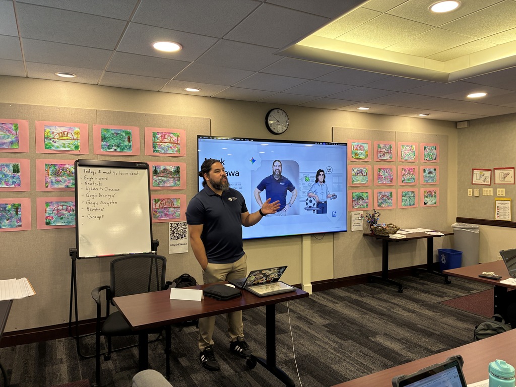 A presenter stands in a board room giving a presentation. He stands next to a whiteboard listing topics like "Google in general" and "Shortcuts," and a large digital screen displaying his name, "Mark," alongside a photo and cartoon avatar of himself.