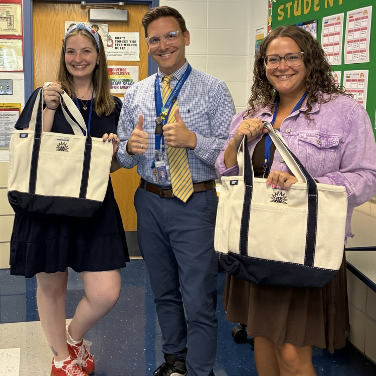 two teachers standing on either side of principal, holding tote bags and smiling in a classroom.
