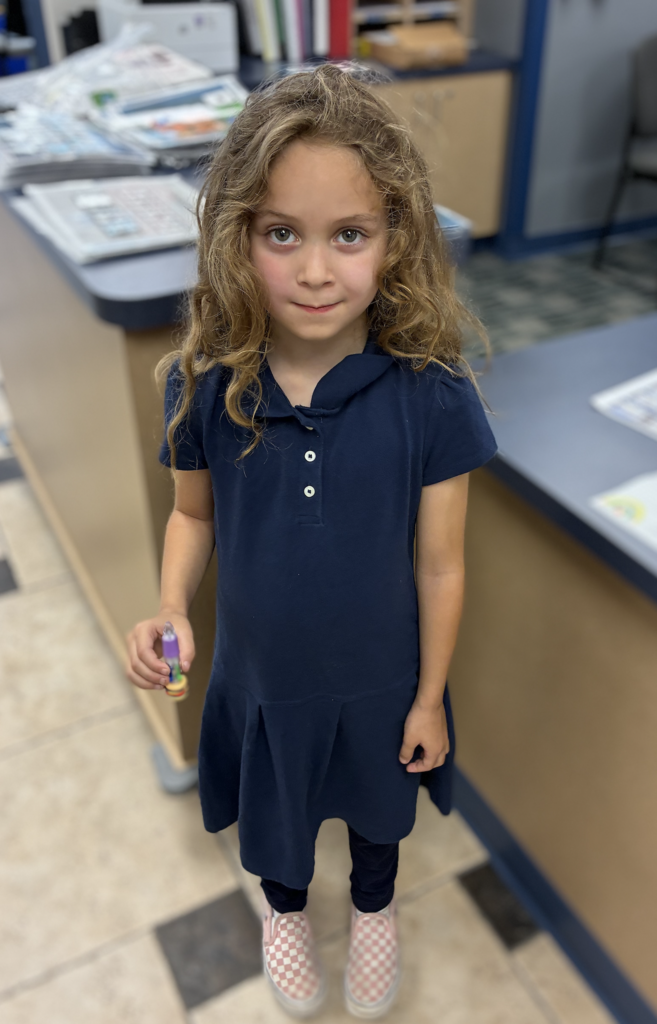 Young girl with wavy blonde hair wearing a navy blue polo dress, dark leggings, and pink checkered slip-on shoes, holding a small toy or novelty pen in a school office.