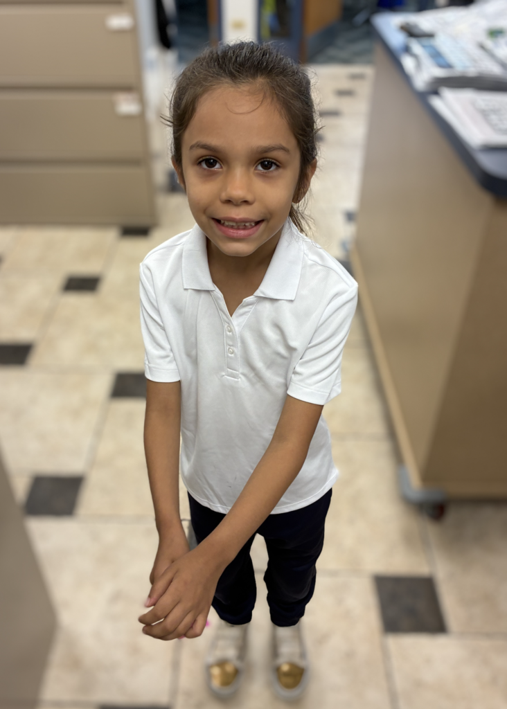 Young girl with brown hair pulled back, wearing a white short-sleeved collared polo shirt, navy pants, and gold-toed shoes, smiling directly at the camera in a school office setting.