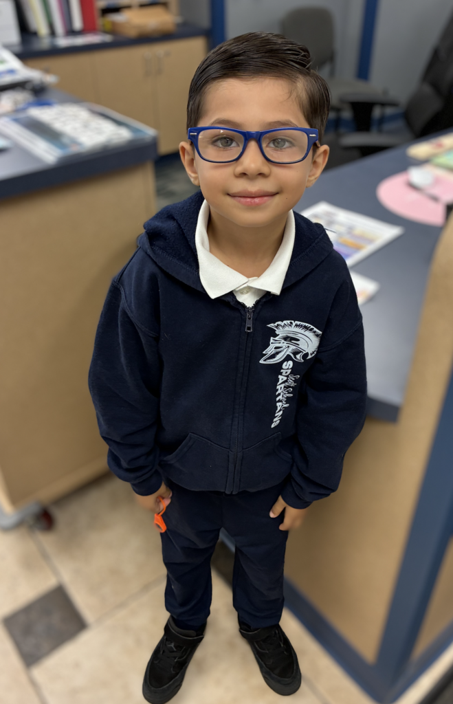 Young boy with blue glasses wearing a navy zip-up hoodie featuring a Spartan mascot logo and the text "Scott School Spartans," standing inside a school office or classroom.