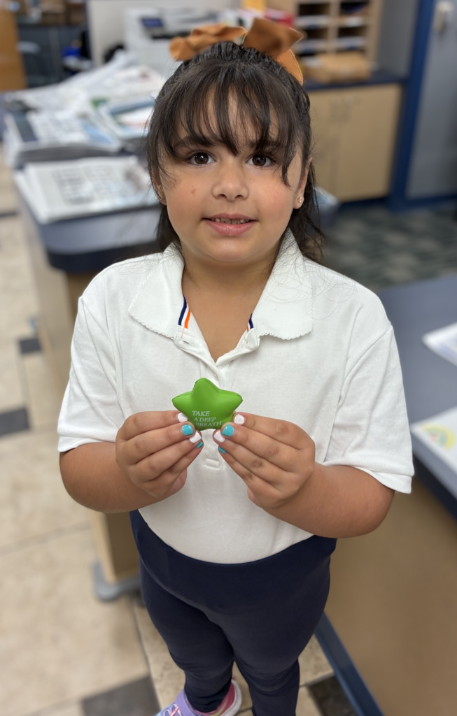 Young girl with bangs and a brown hair bow wearing a white collared polo shirt and navy pants, smiling while holding up a green star-shaped stress ball that reads "TAKE A DEEP BREATH" in a school office.