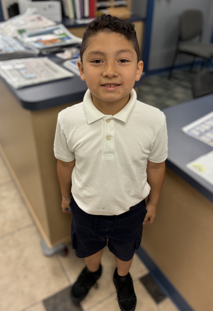 Young boy with dark wavy hair smiling while wearing a white short-sleeved collared polo shirt, navy shorts, and black sneakers, standing in a school office.