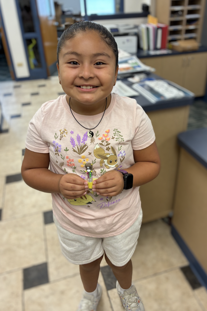 Young girl smiling and holding up a small bear-topped novelty pen inside a classroom or office, wearing a light pink floral Pokémon T-shirt, white shorts, and a yin-yang necklace.