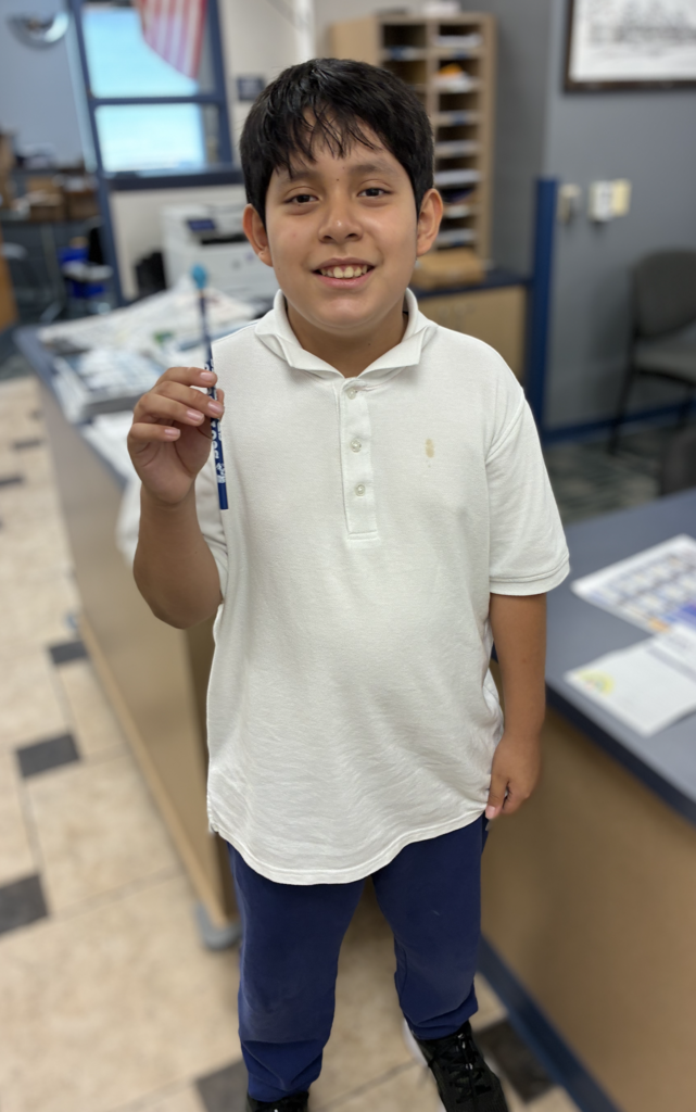 Young boy with dark hair wearing a white collared polo shirt and blue pants, smiling while holding up a blue decorative pencil in a school office.