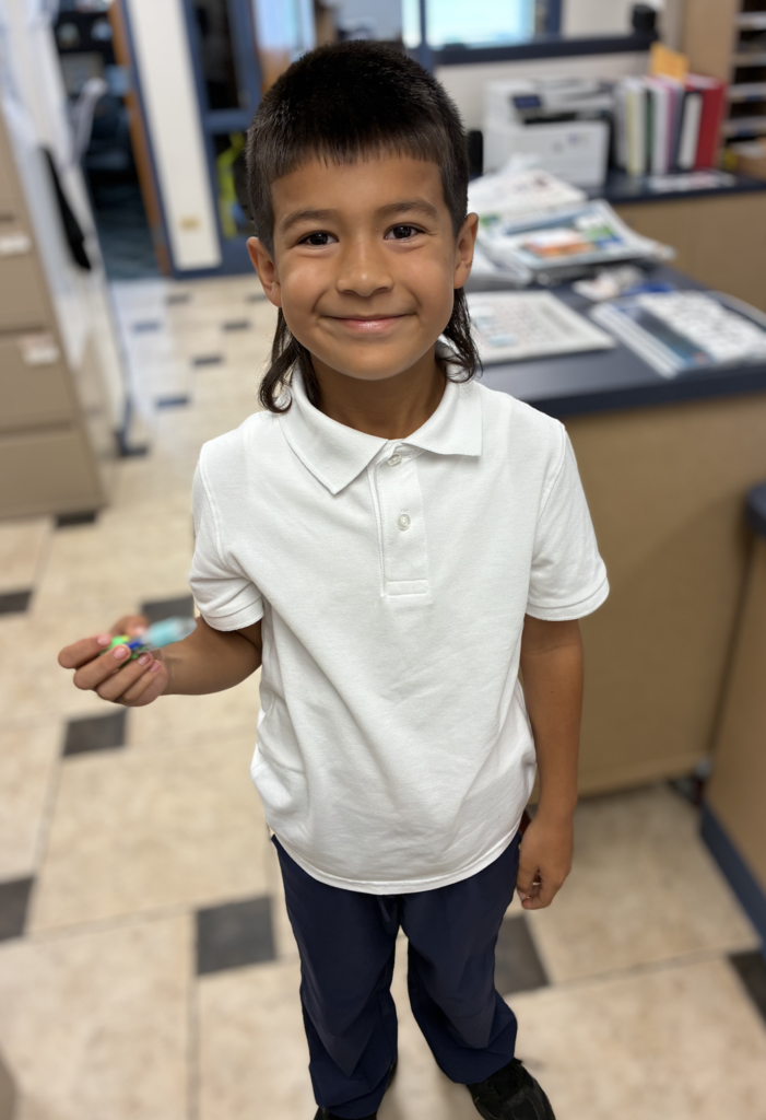 Young boy with dark hair cut in a mullet, smiling while wearing a white short-sleeved polo shirt and navy pants, holding a small toy pen in a school office.