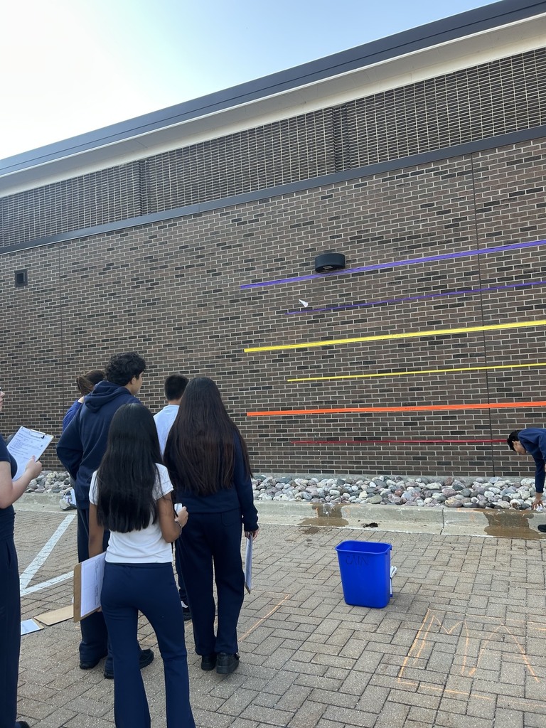A group of school students stands on a paved surface, watching the flight. The brick wall behind the rocket features several horizontal, colored lines in shades of purple, blue, yellow, and orange, which serve as a height or distance scale. A blue plastic bucket sits on the ground near a chalk grid, and clipboard-wielding students track the results.