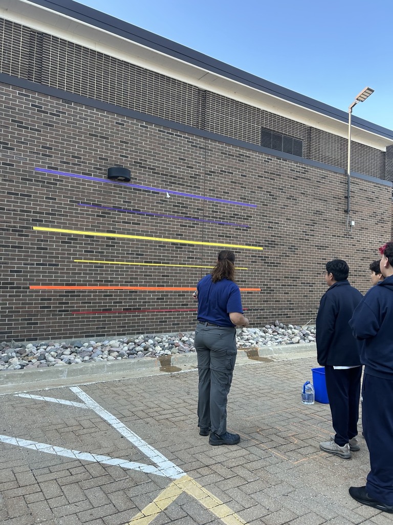 The teacher, seen from behind wearing a blue shirt and grey cargo pants, stands on a paved surface facing a large brick wall. Colorful horizontal lines in purple, blue, yellow, and orange are taped to the wall to measure the rocket's height. A tiny white paper rocket is caught mid-air near the top purple stripe, while a blue bucket and a water jug sit on the ground nearby.
