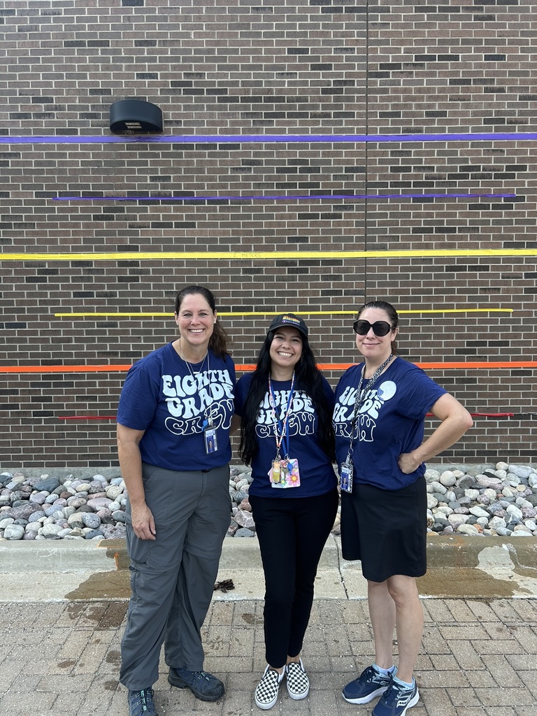 The women stand side-by-side, wearing matching dark blue t-shirts that read "Eighth Grade Crew." The wall behind them features colorful, horizontal vinyl lines in purple, blue, yellow, and orange, with a "2 METERS" label faintly visible on the yellow stripe. They are standing outdoors on a paved area lined with smooth, light-colored landscape rocks.