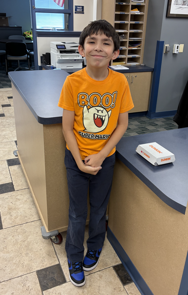 A smiling boy stands in a school front office, wearing a bright orange T-shirt with a Super Mario ghost print that says "BOO!", dark pants, and black and blue sneakers. Next to him on the blue counter sits a small white Dunkin' box.