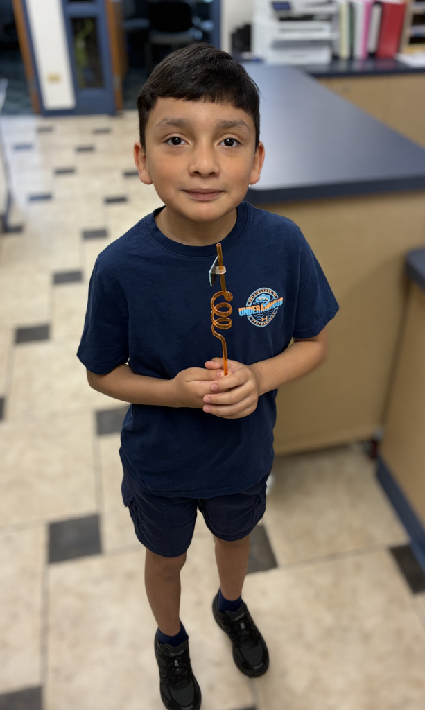 A young boy standing in an office environment, wearing a navy blue T-shirt, matching shorts, and black sneakers, while holding an orange curly straw with both hands.
