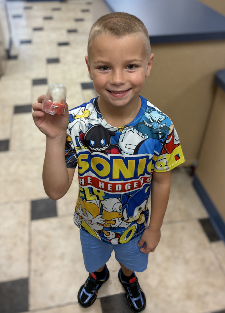 A smiling boy wearing a brightly colored Sonic the Hedgehog T-shirt and blue shorts, holding up a small plastic container in his right hand.