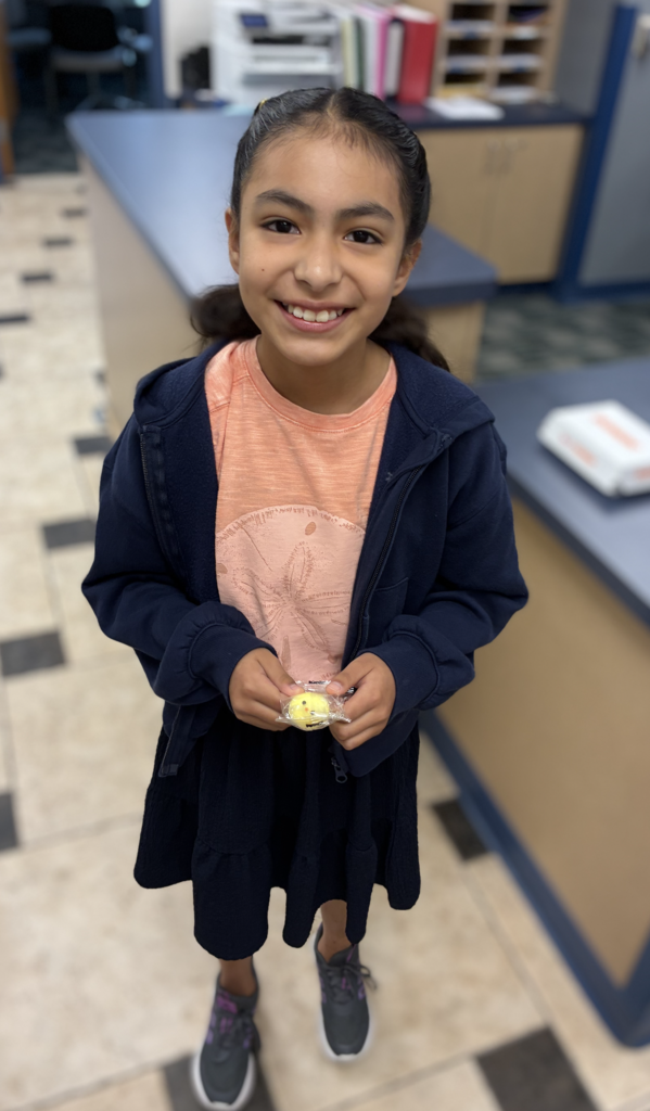 A smiling young girl standing in an office, wearing a dark blue zip-up jacket over a peach t-shirt, a dark blue skirt, and grey sneakers, holding a small wrapped treat in both hands.