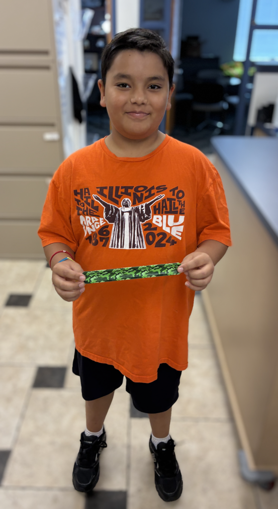 A young boy standing in an office, wearing a bright orange University of Illinois T-shirt and black shorts, holding up a green camouflage slap bracelet with both hands.