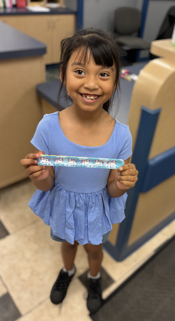 A smiling young girl standing in an office, wearing a light blue peplum top and black sneakers, holding up a blue slap bracelet patterned with white unicorns using both hands.