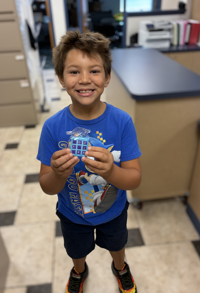 A smiling boy wearing a blue Sonic the Hedgehog T-shirt and dark shorts, holding up a small plastic tic-tac-toe keychain game with both hands.